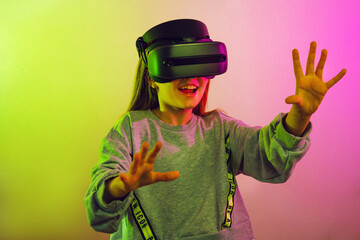 Excited child girl in VR headset touching air during virtual reality experience at studio. Emotional kid exploring artificial computer world or playing video game, on pink background