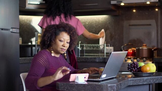 Mother typing in laptop and daughter doing housework. Black woman working and girl cooking in the kitchen.