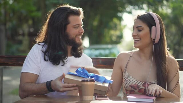 Latin boyfriend giving surprise present to pretty girlfriend sitting outdoors.