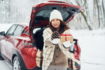 Holding gift box. Beautiful young woman is outdoors near her red automobile at winter time