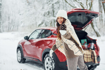 In the forest. Beautiful young woman is outdoors near her red automobile at winter time