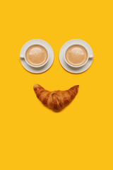 Two cups of coffee with foam on top with a croissant making a smiling happy face for breakfast and morning coffee on a yellow background with copy space and room for text