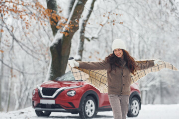 Having a walk in the forest. Beautiful young woman is outdoors near her red automobile at winter time