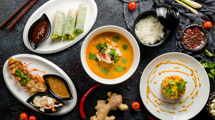 Tom Yam soup, spring rolls with shrimp, boiled rice, tartar from salmon and fried pork with sauce top view on dark stone table, Chinese cuisine