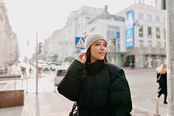 Adorable lovely girl with big green eyes and nude make up wearing black jacket and beige cap looking aside and smiling on blurred city background