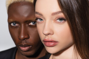 close up view of brunette woman looking at camera near blurred african american man isolated on grey.