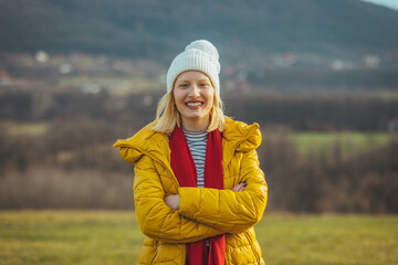 Portrait of an attractive young woman standing outside in a field. she is dressed in warm clothes and looking at the camera. She's smiling .