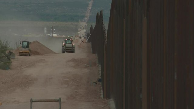 A Construction Truck Carrying Pipes Drives Towards Camera Along The Newly Constructed Border Wall Between US And Mexico