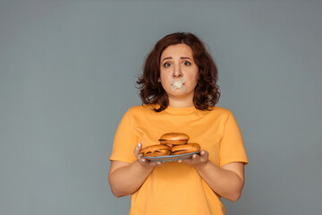 giving up fast food. upset girl with her mouth sealed with adhesive tape holds a plate with burgers in her hands. photo on a gray background