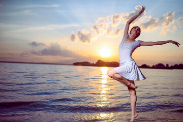Fototapeta na wymiar Love and Passion To Dance With Japanese Ballet Dancer in White Dress And Silver Crown Standing In Ocean Wave And Showing Ballet Pas With Lifted Hands Against Picturesque Sunset At Sea.