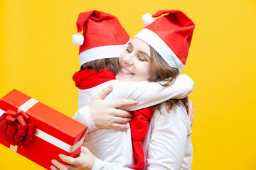 Portrait of Two Beautiful Caucasian Ladies of Different Ages Embracing Together in Santa Hats With Wrapped Red Giftbox Behind and Casual Clothes Against Trendy Yellow Background. Vertical Image