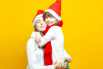 Obraz premium Two Caucasian Blond Sisters in Winter Santa Hats Embracing Cuddling Together in White Casual Clothing Over Yellow Background