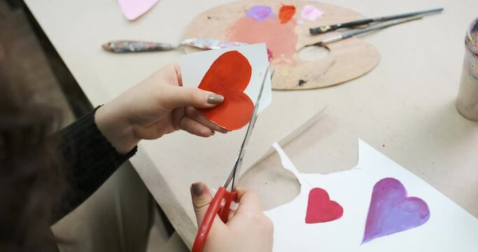 Cutting a card for St Valentine's Day from paper. Painted handmade red heart as a gift. Crafted homemade present, preparing for celebrating holiday.