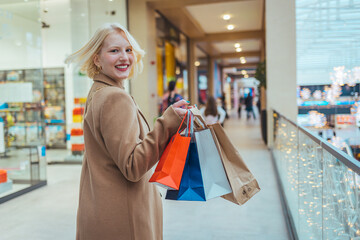 Young blonde woman walking back to camera in shopping center on the background of boutiques. Beauty woman in a denim jacket with shopping bags in shopping mall. Copy space