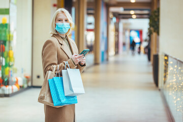 Portrait of casually dressed confident young woman wearing protective medical mask while walking in mall with bunch of shopping bags in hands. Black friday sales concept.