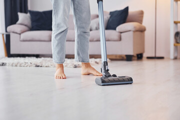 Close up view of legs and vacuum cleaner. Young woman in casual clothes is indoors in domestic room