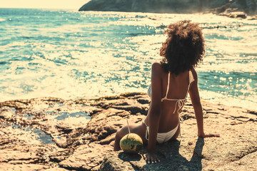 View from behind of a ravishing young African-American woman with curly African hair tanning while sitting on the rock of a beach and enjoying dazzling sunlight with a teal ocean and coco next to her - obrazy, fototapety, plakaty