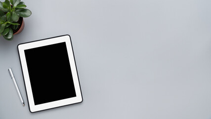 The top view image of a gray workspace is surrounded by a white tablet with a blank screen and a flower in a pot.