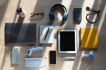 Office desk in sunlight and rainbow backlight. Office workplace mockup with office equipment and stationery and COVID -19 protection and prevention tools. Computer, tablet, smartphone, headphones