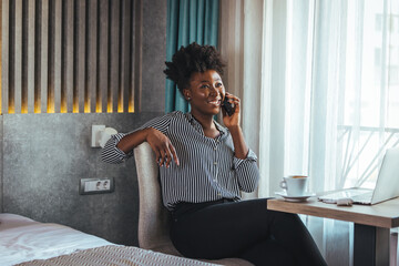 A happy business woman is talking on a mobile phone while sitting at the table of a hotel room with...