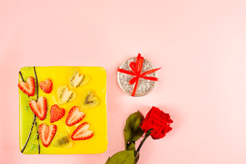 appetizing cake covered with icing, decorated with hearts of strawberries and kiwi pieces with a rose and a gift box on a light background, flat lay. Greeting card. Cake for Valentine's Day.