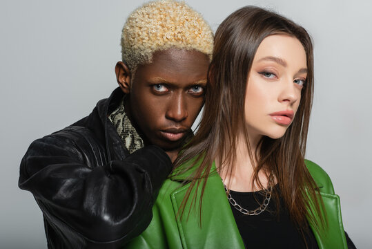 brunette woman and blonde african american man in leather jackets looking at camera isolated on grey.