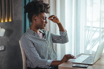 Young frustrated woman working at desk in front of laptop suffering from chronic daily headaches,...