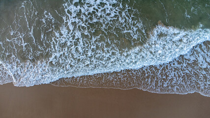 Aerial drone view perpendicular to the blue waves crashing on the sandy beach at the coast. The impact with wet wax creates white foam. The winter beach is empty. High quality photo