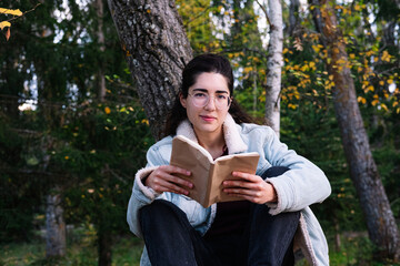 A young woman with glasses reading a book sitting on a tree branch. Reading concept.