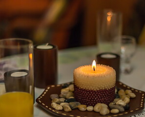 Lit decorative candle on a saucer with stones on the background of candles and glasses on the table