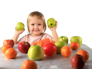 Fototapeta na wymiar A 3-year-old girl holds green apples in her hands and there are a lot of fruits on the table: orange oranges, tangerines, red apples and persimmons as an option for healthy nutrition of children 