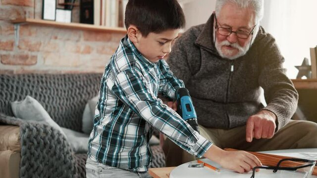 Old grandfather shows and teaches the little boy how to use a electrical drill when assembling furniture. Skills carpentry.