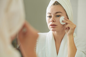 Womam in bathrobe looking at mirror and using cotton pad when applying toner on face