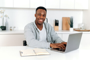 Obraz na płótnie Canvas Friendly black man, freelancer, designer or student, sitting at home in the kitchen, using laptop and notebook study or surf the Internet, improving his skills, smiling friendly. E-learning concept