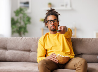 Bored tired african american man changing tv channel with remote control while relaxing at home