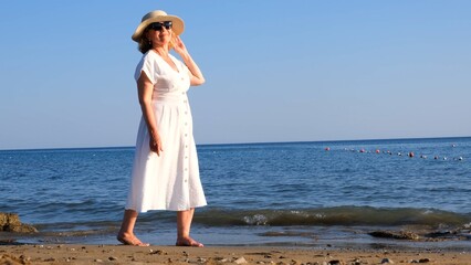 Fototapeta na wymiar mature woman in sunglasses in a straw hat and white dress walks along the blue sea coast on a sunny summer day, enjoying freedom and relaxation. The concept of a senior citizen everyday life