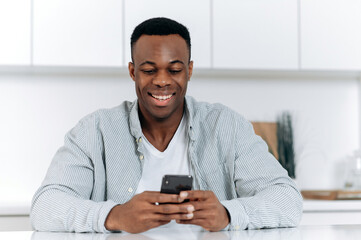 Obraz na płótnie Canvas African American attractive positive young man, in stylish casual clothes, spend leisure time sitting at the table in the kitchen uses smartphone, browses internet, chatting in social media, smiles