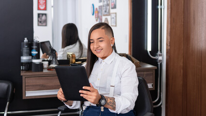 Happy professional barber woman looking at tablet at barber shop.