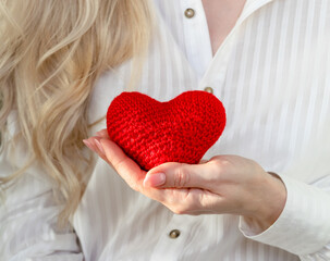 A blonde woman in a white blouse holds a red knitted heart in her hand. The concept of care, love and Valentine's day. Cardiovascular diseases.