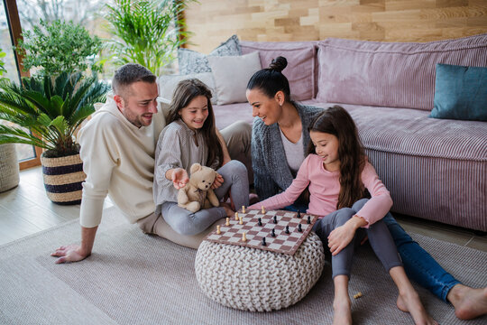 Two happy sisters with mother and father sitting on floor and playing chess together at home