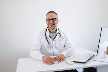 Portrait of smiling doctor in eyeglasses and in white coat sitting at his workplace at office