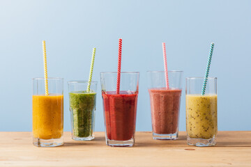 Set of multi-colored fruit smoothies in glasses with paper tubes on wooden table and blue background. Mango, kiwi, strawberry, apple summer drinks