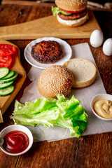 Obraz na płótnie Canvas Cooking hamburger or cheeseburger. Different ingredients for a classic hamburger. Grilled meat, vegetables, greens, sauces near a sesame bun. Wooden background.