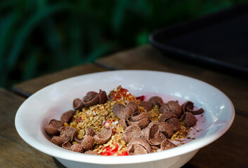 a bowl of strawberry yogurt with granola, fruit and chocolate cornflakes on a wooden table.