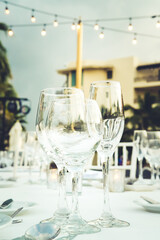 Festive serving with glasses for an outdoor celebration with garland lamps and palms at the background. Wedding, anniversary, birthday dinner and party. Toned image with place for text