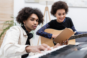 Smiling african american woman playing acoustic guitar near boyfriend with synthesizer and notebook at home.