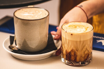 Couple of cappuccino with foam served by the hand of a girl at a bar counter, one in a ceramic mug, one in a glass