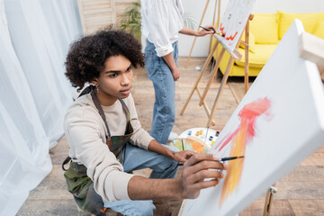 African american man in apron painting on blurred canvas near girlfriend at home.