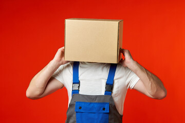 Man in work clothes got his head stuck in a cardboard box and is trying to take it off on a red background. Misunderstanding during work. Photo