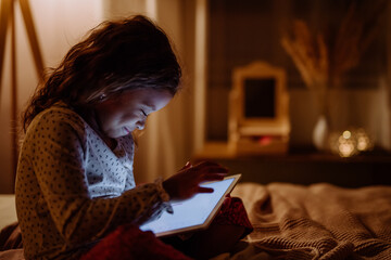 Fototapeta na wymiar Happy little multiracial girl using tablet on bed in evening at home.